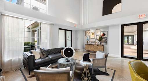 A black leather couch sits in a brightly lit room with a yellow chair and a table with a fan.
