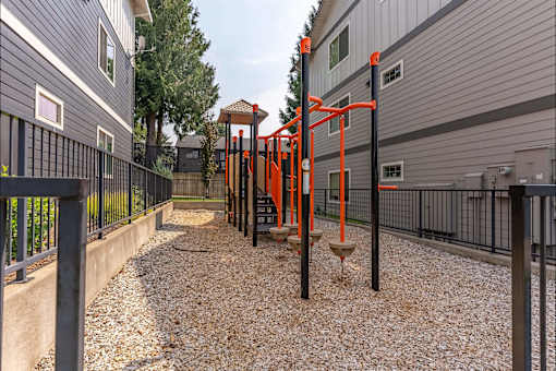 A playground with orange and black equipment is situated between two grey buildings at Sandyplace Apartment Homes, Sandy, OR