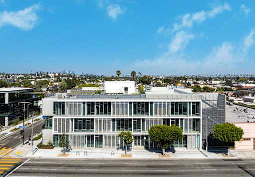 A modern building with a glass facade is situated on a street corner with trees and other buildings in the background.