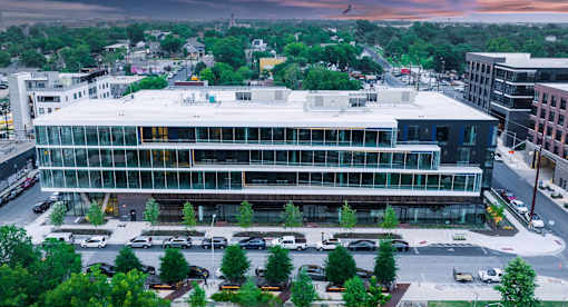 A modern glass and steel building is surrounded by trees and parking.