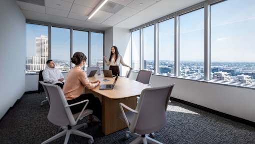 A woman is giving a presentation to two people at a conference table in a room with a city view.