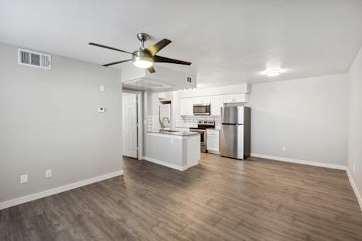 A room with a ceiling fan and a kitchen area in the background.