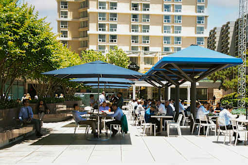 People are sitting at tables under blue umbrellas in a courtyard.