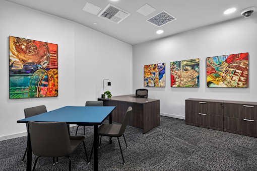 A conference room with a blue table and chairs, a desk with a chair, and three paintings on the wall.