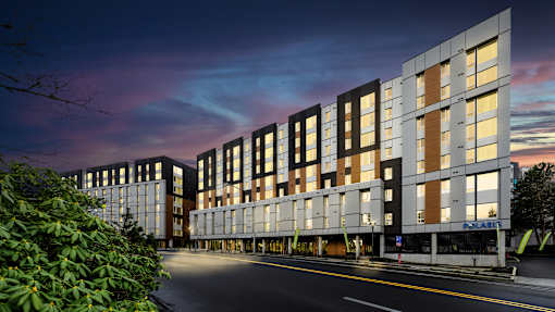 A modern building with a glass facade is illuminated from within, with a tree to the left and a street in front.
