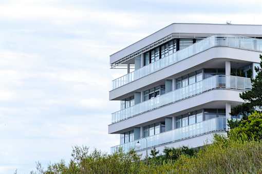 A white building with balconies is surrounded by greenery.