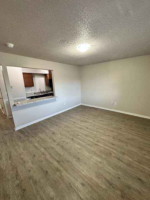 A room with a wooden floor and a kitchen visible through a glass door.