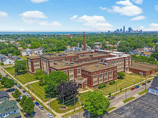 A large red brick building with a tall chimney is surrounded by green trees and grassy areas.