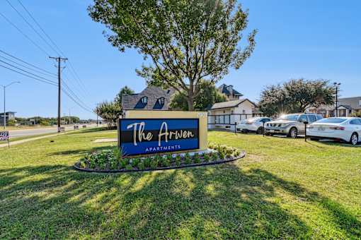 A sign for "The Arrow Apartments" is displayed in front of a grassy area with a tree and parked cars.
