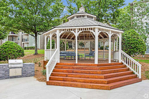 A gazebo with a wooden deck and stairs is surrounded by green trees.