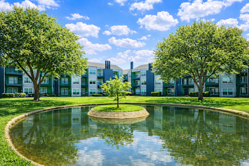 A small pond in the middle of a grassy area with trees and apartment buildings in the background. at Infinity on the Landing Apartments , Texas, 76040