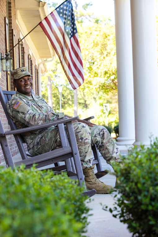 A soldier in uniform sits on a porch with an American flag in the background.