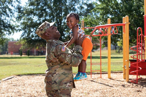 A soldier in camouflage uniform is lifting a young girl in a playground.