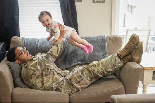A man in military uniform is lying on a couch with a baby on his feet.