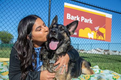 A woman is hugging a German Shepherd dog in front of a sign that says Bark Park.