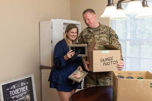 A man in military uniform is holding a box labeled "Family Photos" while a woman in a blue dress looks on.