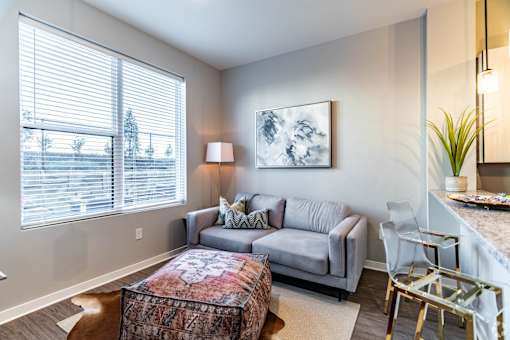 A living room with a grey couch, a patterned ottoman, and a painting on the wall.
