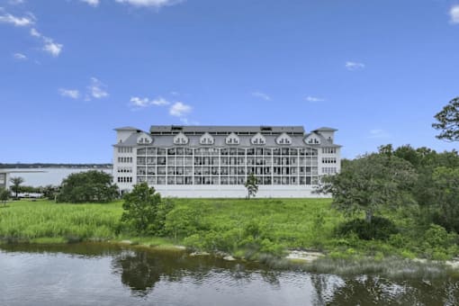 A large white building with a grey roof is surrounded by greenery and water.