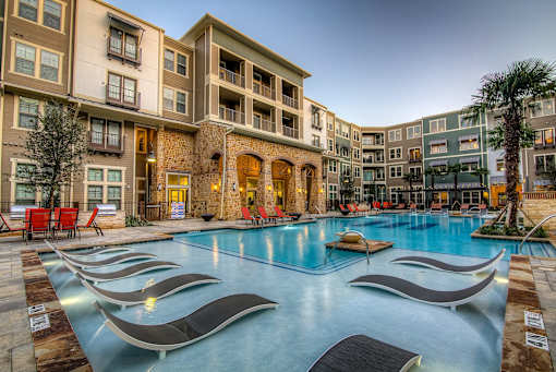 A large swimming pool surrounded by lounge chairs in front of apartment buildings.