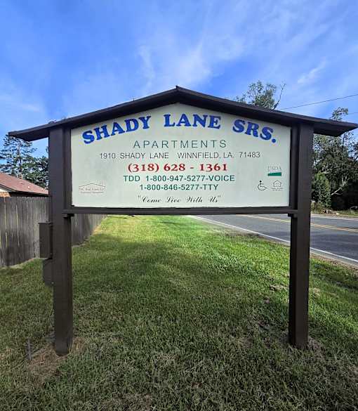 A sign for Shady Lane SRS Apartments is displayed in front of a grassy area.