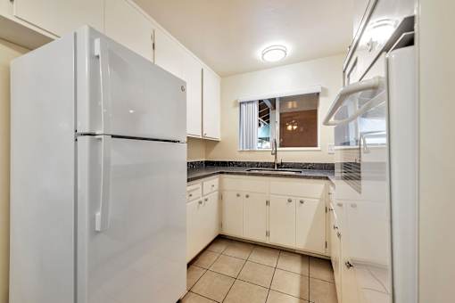 A white refrigerator stands in a kitchen with white cabinets and a tiled floor.