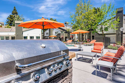 A silver grill is in the foreground of a patio with orange chairs and umbrellas.