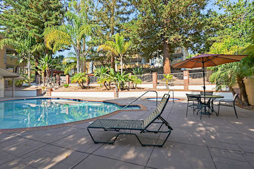 A pool surrounded by trees and a patio with chairs and an umbrella.