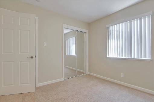A white door is on the left of a room with a carpeted floor and a window with blinds.