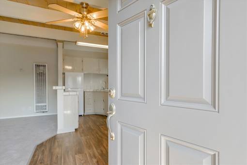 A white door with a gold handle is open to a room with a ceiling fan and a kitchen in the background.
