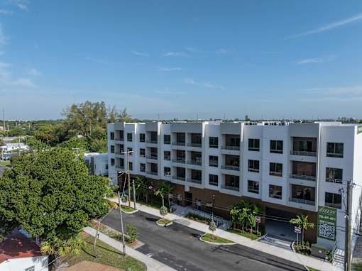A large white building with balconies is surrounded by trees and has a parking lot in front.