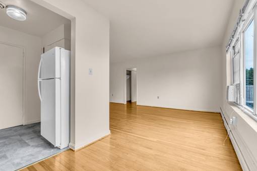 A white refrigerator is in the corner of a room with wooden floors and white walls.
