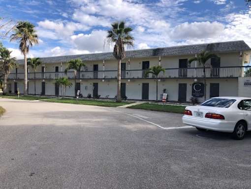 A white car is parked in a parking lot in front of a building with palm trees.