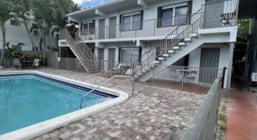 A pool is surrounded by a brick patio and a building with a balcony and stairs.
