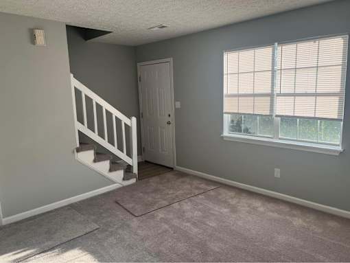 A room with a carpeted floor, a staircase, and a window with blinds.