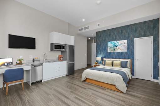 A modern bedroom with a bed, desk, and kitchenette.