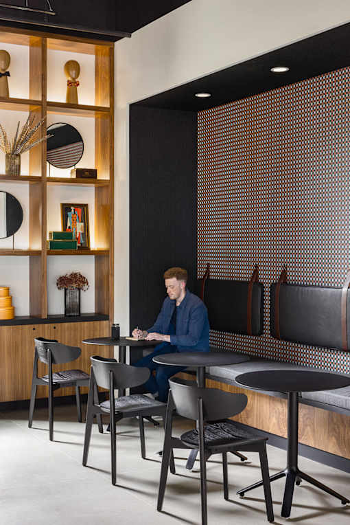 A man is sitting at a table in a room with a wooden shelf and a black and white patterned wall.