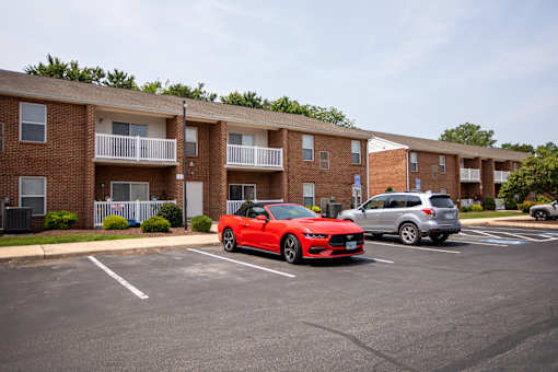 A red sports car is parked in a parking lot in front of a brick apartment building.