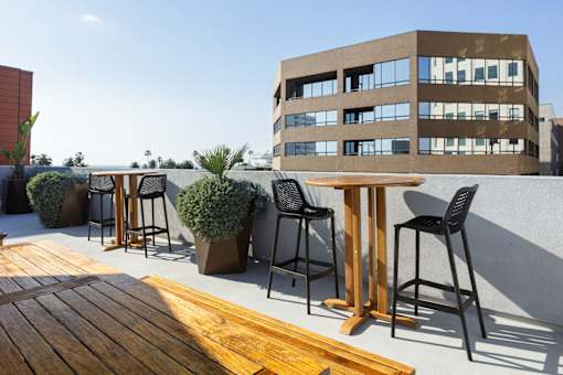 A wooden table is in the foreground of a patio with black chairs and a building in the background.