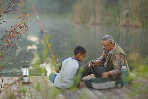 A man and a boy are sitting by a body of water, with the man holding a fishing rod.