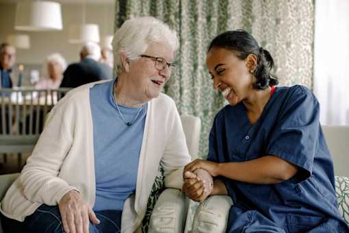 An elderly woman in a blue shirt and a young woman in a blue scrub are holding hands.