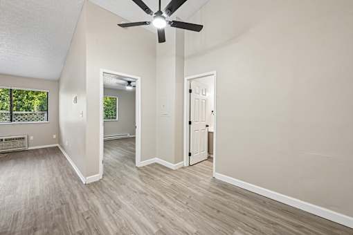 A room with a ceiling fan and light fixture, hardwood floors, and a doorway leading to another room.