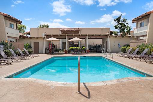 A large swimming pool in front of a house with a patio and lounge chairs.