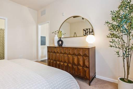 A bedroom with a white bed, a wooden chest of drawers, a round mirror, and a potted plant.