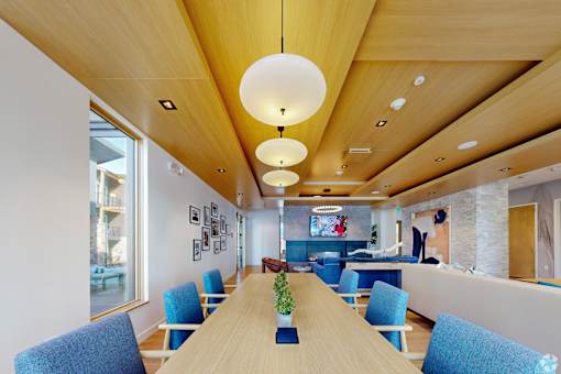 A long wooden table with blue chairs is in the middle of a room with a white wall and a TV.