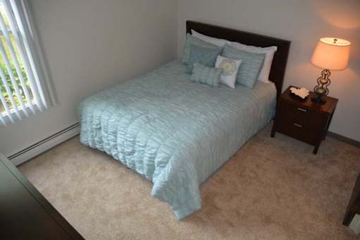 A bed with a blue comforter and two pillows sits in a room with a lamp and a nightstand.