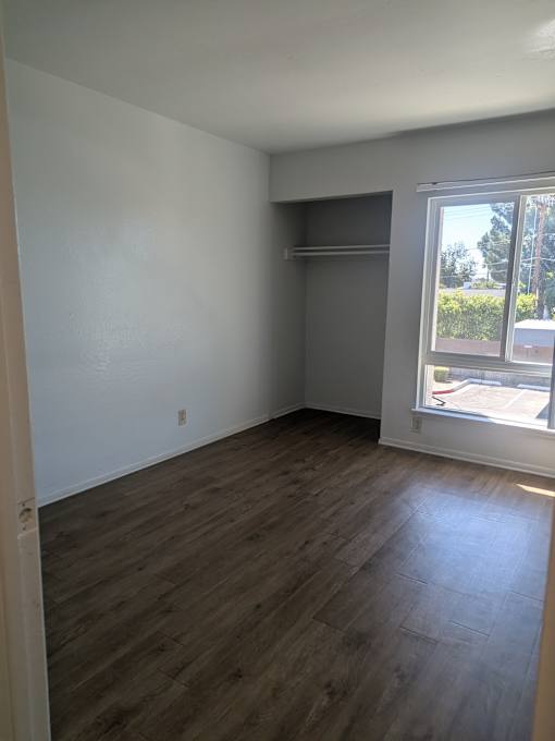 A room with wooden flooring and a window showing a view of a parking lot and trees.