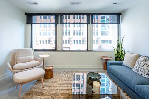 A living room with a blue couch, a chair, a small table, and a window with a view of a city.
