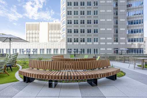A wooden bench sits in the middle of a paved area with a grassy area and a building in the background.