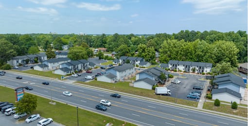 A road with cars and a parking lot with cars and houses in the background.