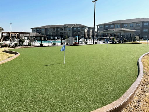 an artificial grass putting green with a pool and apartment buildings in the background